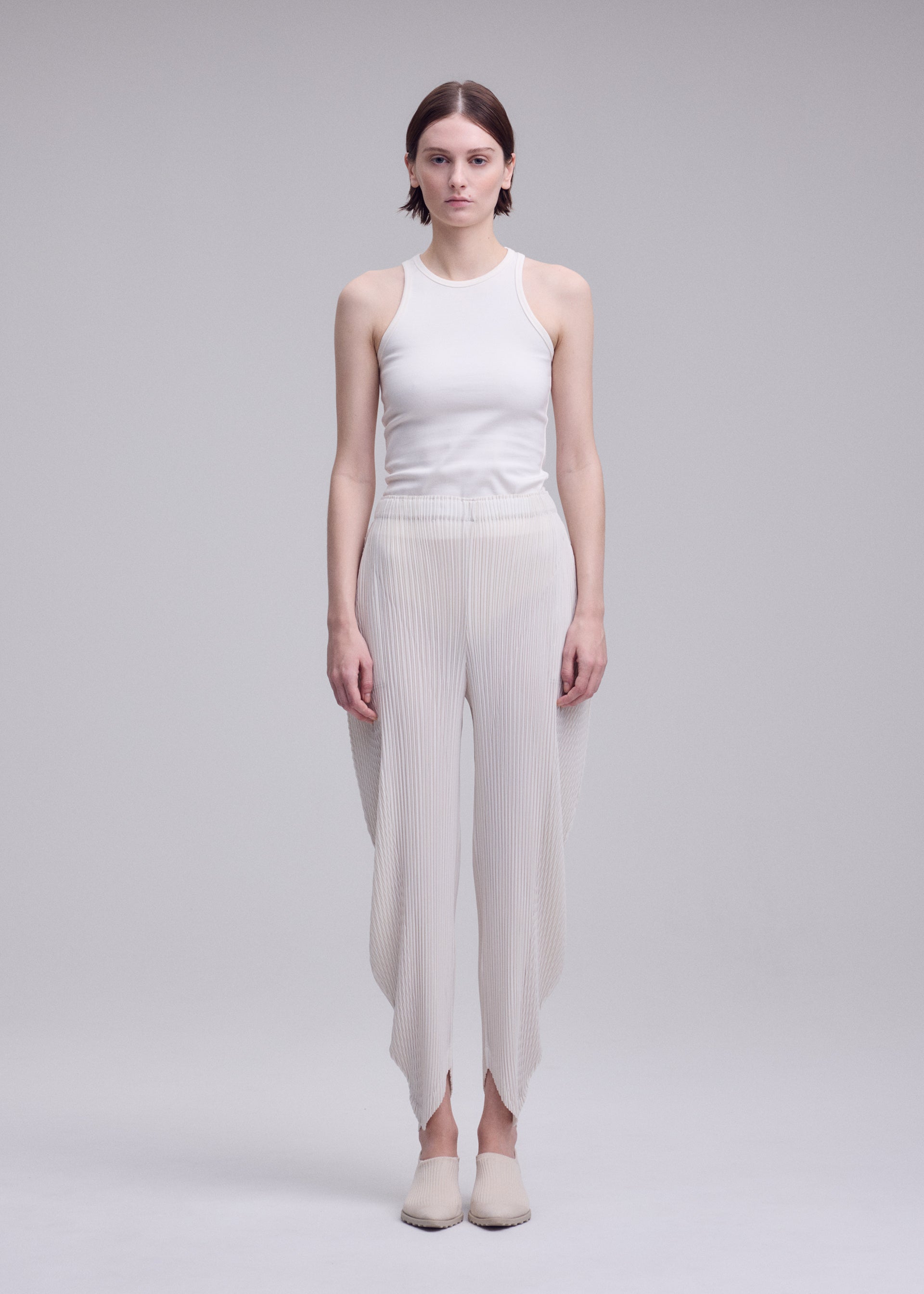 Zoomed view A woman stands facing forward against a plain light gray background, wearing the ISSEY MIYAKE LEAF PLEATS: a sleeveless white top paired with double-pleated, asymmetrical pants, and flat shoes.