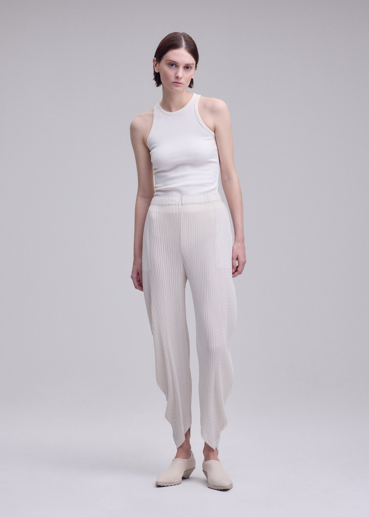 A person with short brown hair and a neutral expression stands against a plain background wearing ISSEY MIYAKE’s LEAF PLEATS textured double-pleated pants with wavy hems, paired with a white sleeveless top and beige slippers. view 7 of 8