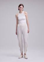 A person with short brown hair and a neutral expression stands against a plain background wearing ISSEY MIYAKE’s LEAF PLEATS textured double-pleated pants with wavy hems, paired with a white sleeveless top and beige slippers.