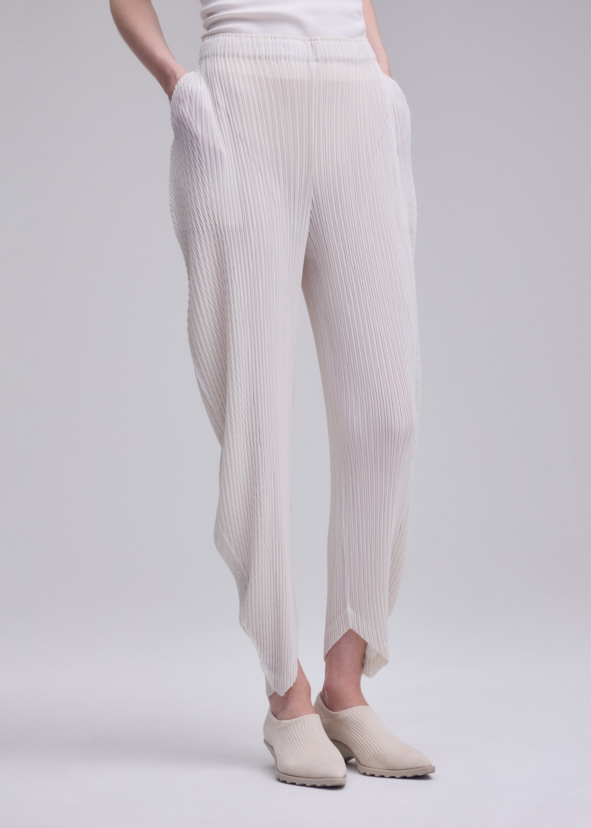 Zoomed view Dressed in ISSEY MIYAKE’s LEAF PLEATS—crisp white double-pleated pants with an asymmetrical hem and subtle leaf silhouettes—this individual pairs the elegant trousers with matching slip-on shoes, posing against a plain light background with hands comfortably in their pockets.