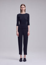 A person faces forward in a sleek silhouette, wearing ISSEY MIYAKE's RHOMBUS PLEATS black fitted top with three-quarter sleeves and matching ankle-length pants, paired with black pointed shoes, against a plain light gray background.