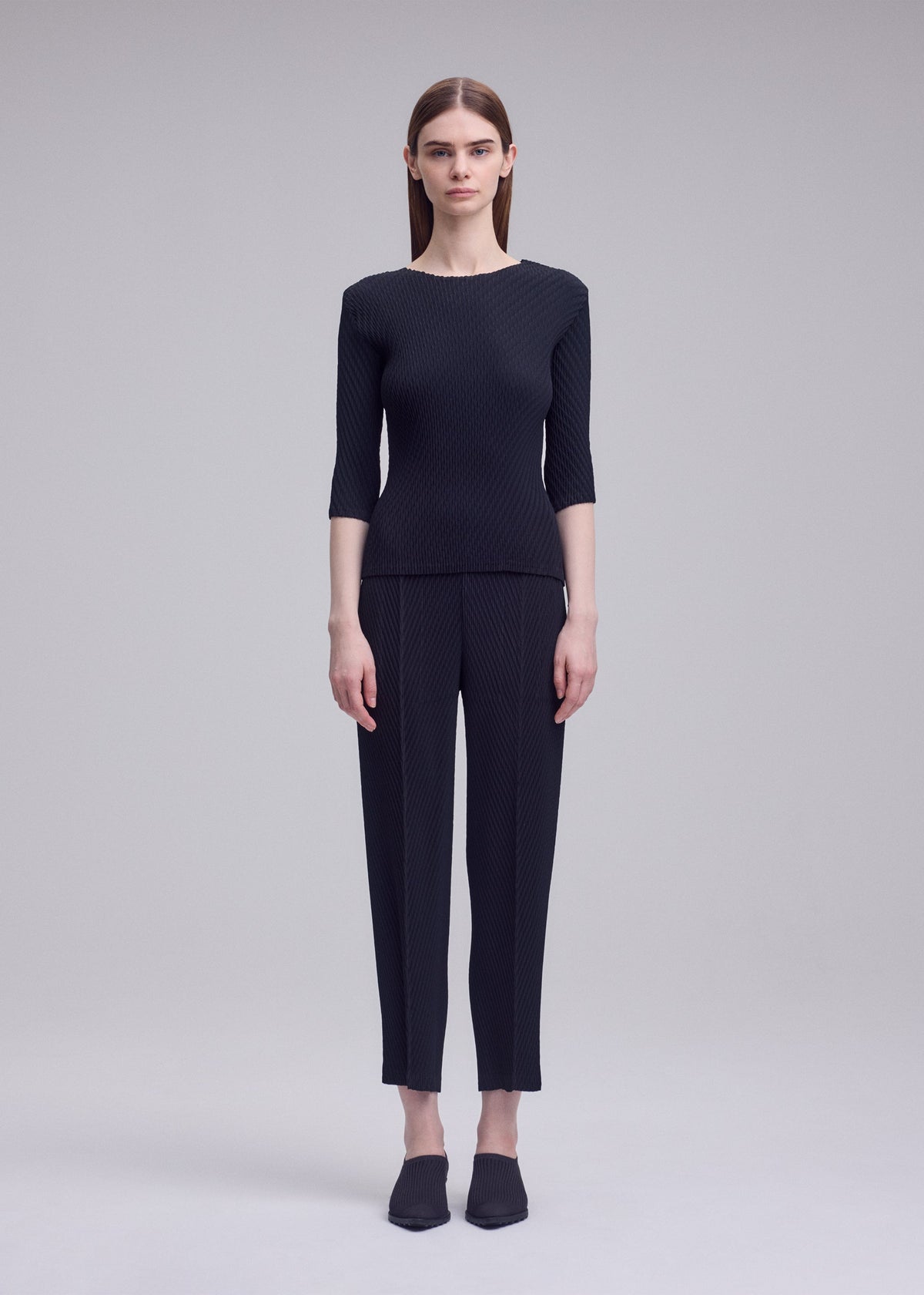 A person with straight brown hair stands against a gray background, wearing ISSEY MIYAKE's RHOMBUS PLEATS: a sleek black top and ankle-length pants, paired with black shoes. view 3 of 5