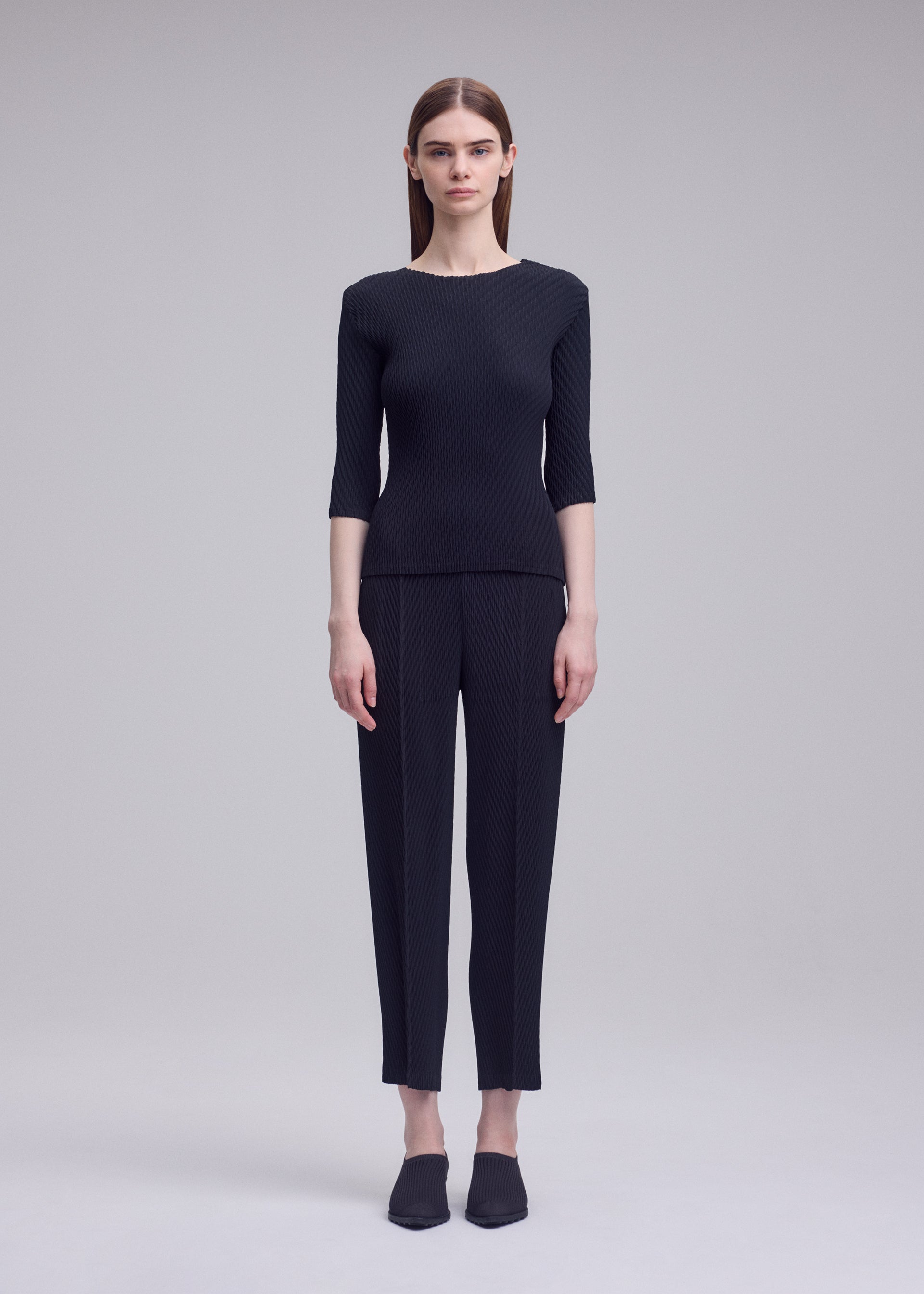 Zoomed view A person with straight brown hair stands against a gray background, wearing ISSEY MIYAKE's RHOMBUS PLEATS: a sleek black top and ankle-length pants, paired with black shoes.