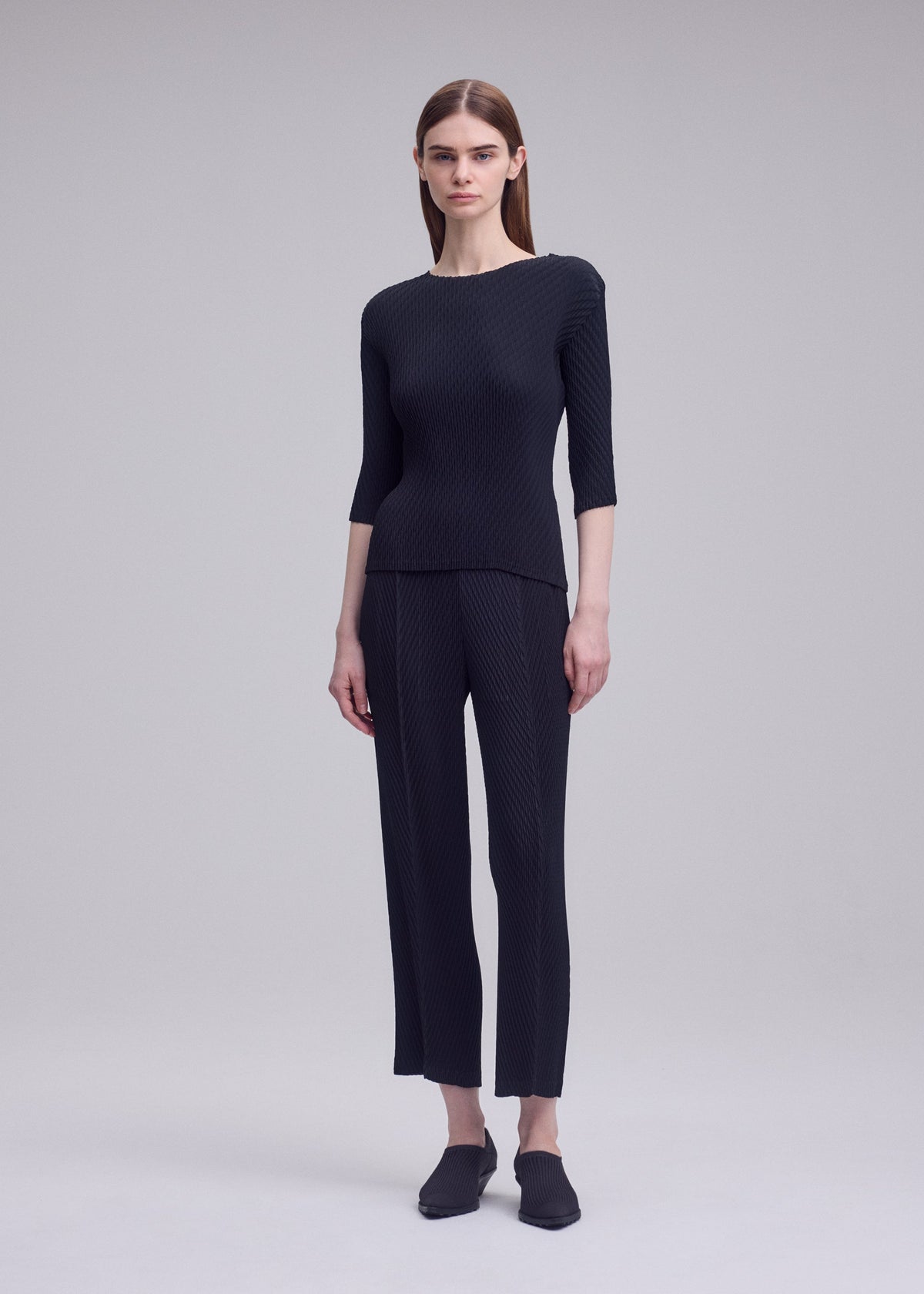 A person with straight brown hair stands against a plain grey background, wearing ISSEY MIYAKE's RHOMBUS PLEATS fitted dark textured top with three-quarter sleeves and matching double-pleated straight-leg pants. view 5 of 6