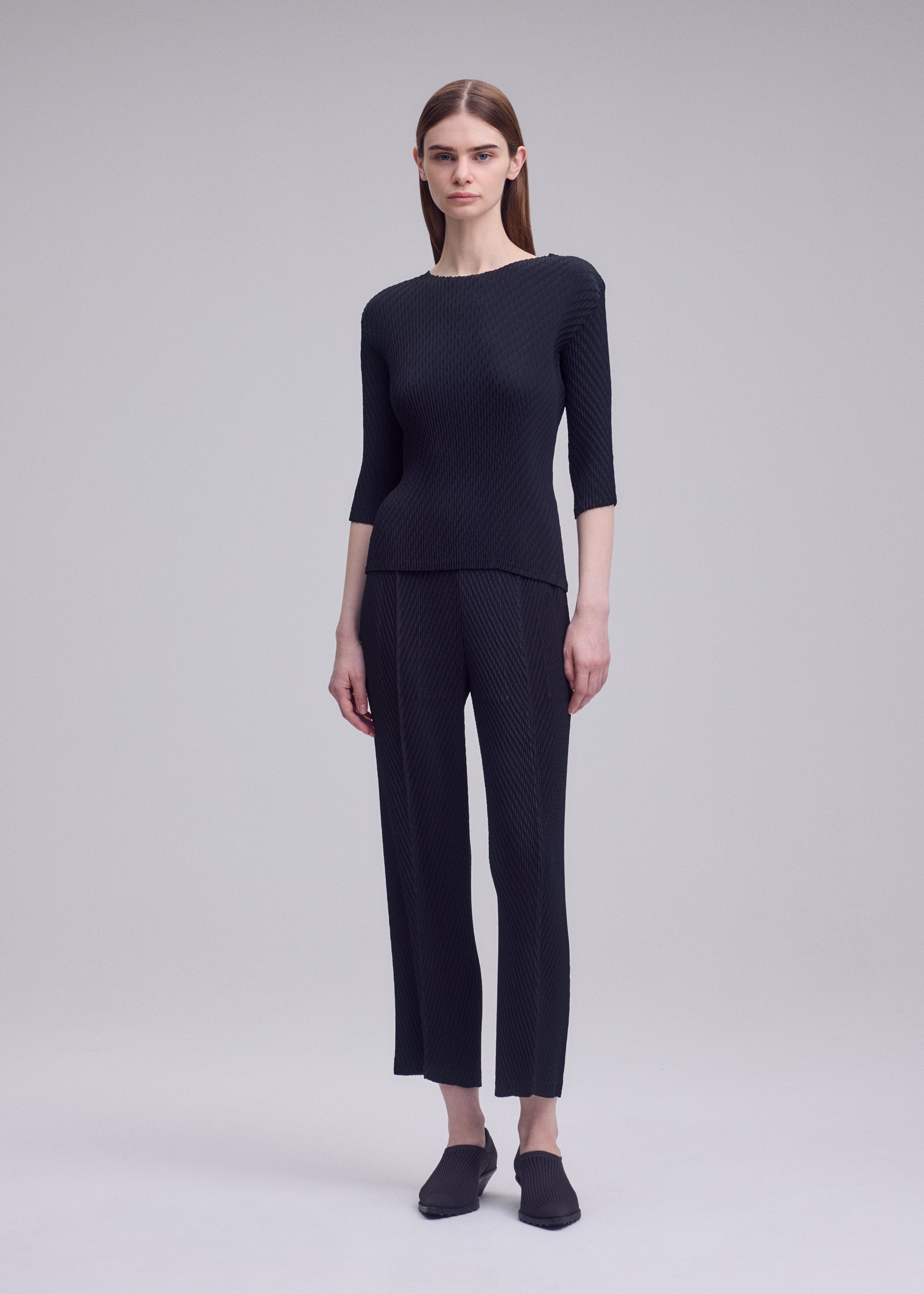 Zoomed view A person with straight brown hair stands against a plain grey background, wearing ISSEY MIYAKE's RHOMBUS PLEATS fitted dark textured top with three-quarter sleeves and matching double-pleated straight-leg pants.