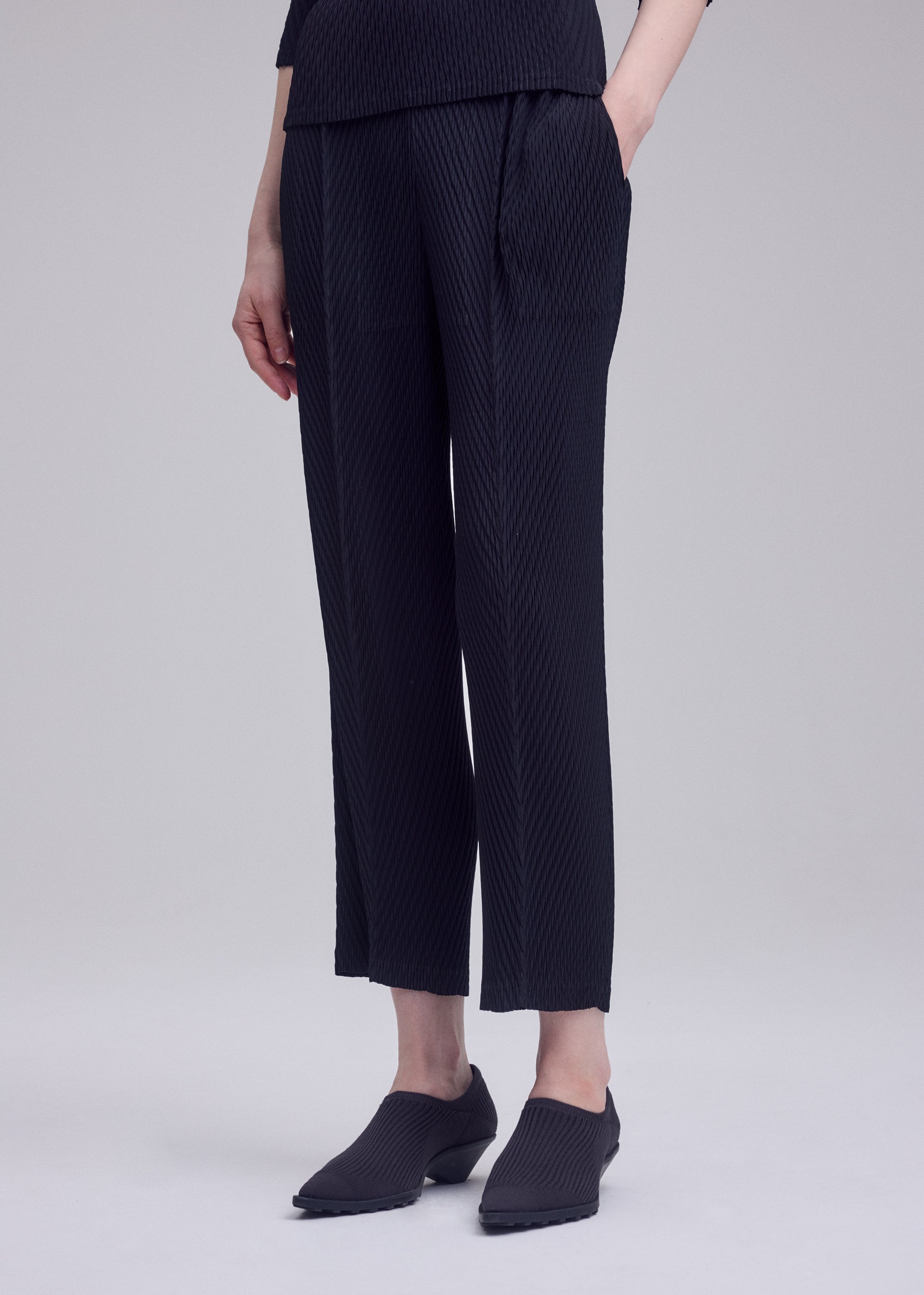 Zoomed view A person stands with one hand in their pocket, wearing ISSEY MIYAKE RHOMBUS PLEATS black textured straight-leg pants in a relaxed fit and black slip-on shoes, set against a plain light gray background.