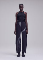 A person with braided hair wears the ISSEY MIYAKE HANDS OF LOVER PLEATS: a black tank top paired with loose black pleated pants adorned with a distinctive white original pattern, inkjet-printed on soft polyester crepe de chine. They stand against a plain, light gray background with a neutral expression.