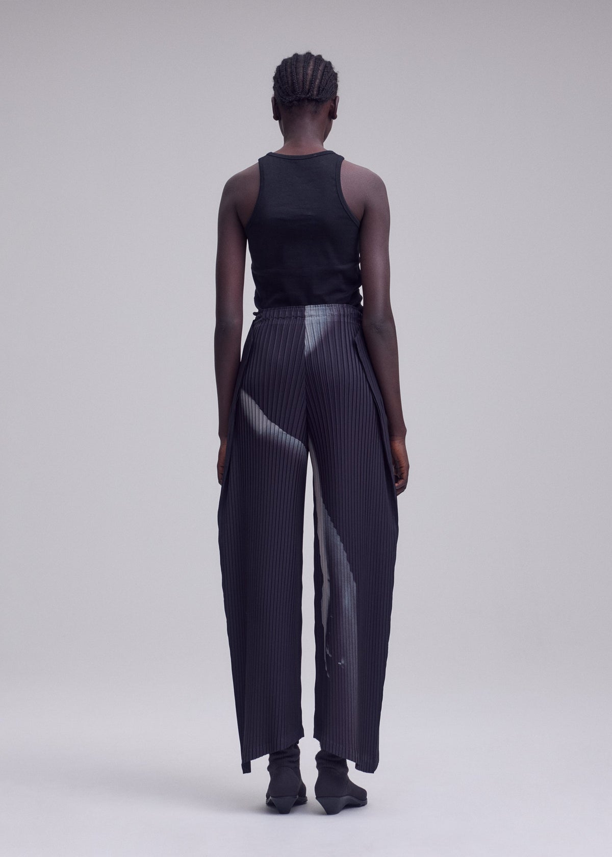 A person with short, braided hair stands facing away, wearing a black tank top and ISSEY MIYAKE’s HANDS OF LOVER PLEATS—high-waisted, wide-leg pants in black polyester crepe de chine featuring a subtle original white pattern—set against a plain, light gray background. view 6 of 7
