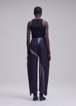 A person with short, braided hair stands facing away, wearing a black tank top and ISSEY MIYAKE’s HANDS OF LOVER PLEATS—high-waisted, wide-leg pants in black polyester crepe de chine featuring a subtle original white pattern—set against a plain, light gray background.