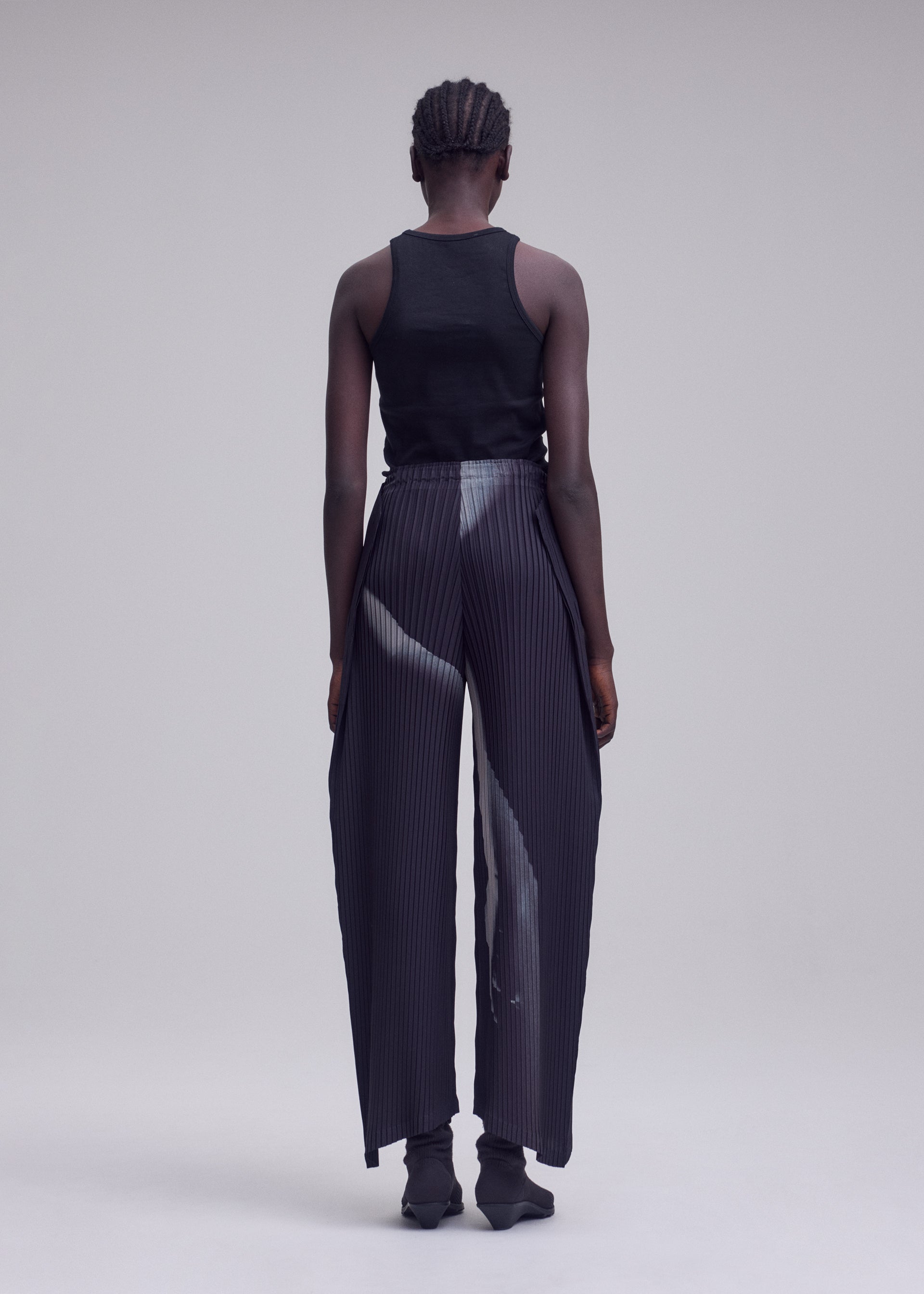 Zoomed view A person with short, braided hair stands facing away, wearing a black tank top and ISSEY MIYAKE’s HANDS OF LOVER PLEATS—high-waisted, wide-leg pants in black polyester crepe de chine featuring a subtle original white pattern—set against a plain, light gray background.