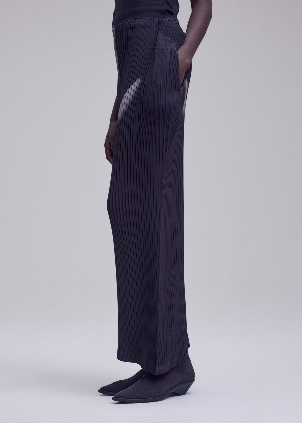 A person stands against a plain light background with one hand in their pocket, dressed in ISSEY MIYAKE’s HANDS OF LOVER PLEATS—black wide-leg pants crafted from polyester crepe de chine—paired with a black top and black shoes. view 7 of 7