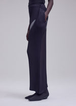 A person stands against a plain light background with one hand in their pocket, dressed in ISSEY MIYAKE’s HANDS OF LOVER PLEATS—black wide-leg pants crafted from polyester crepe de chine—paired with a black top and black shoes.
