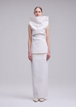 A woman stands against a plain background wearing ALLOW by ISSEY MIYAKE—a minimalist, structured white washi paper-blend outfit with a high, wide collar and long skirt. Her hair is pulled back; her arms rest relaxed by her sides.