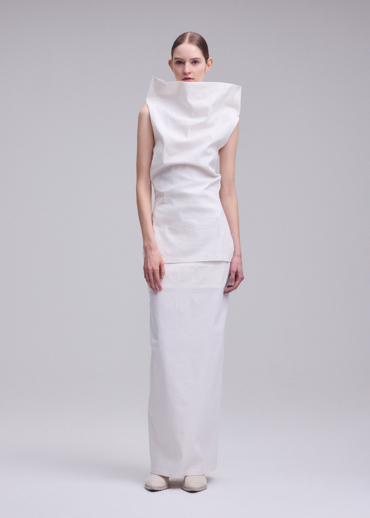 A woman stands against a plain background in the ALLOW dress by ISSEY MIYAKE—a structured, high-collared white piece with a long washi paper-blend skirt. She completes her modern minimalist look with minimal accessories and white shoes. view 7 of 9