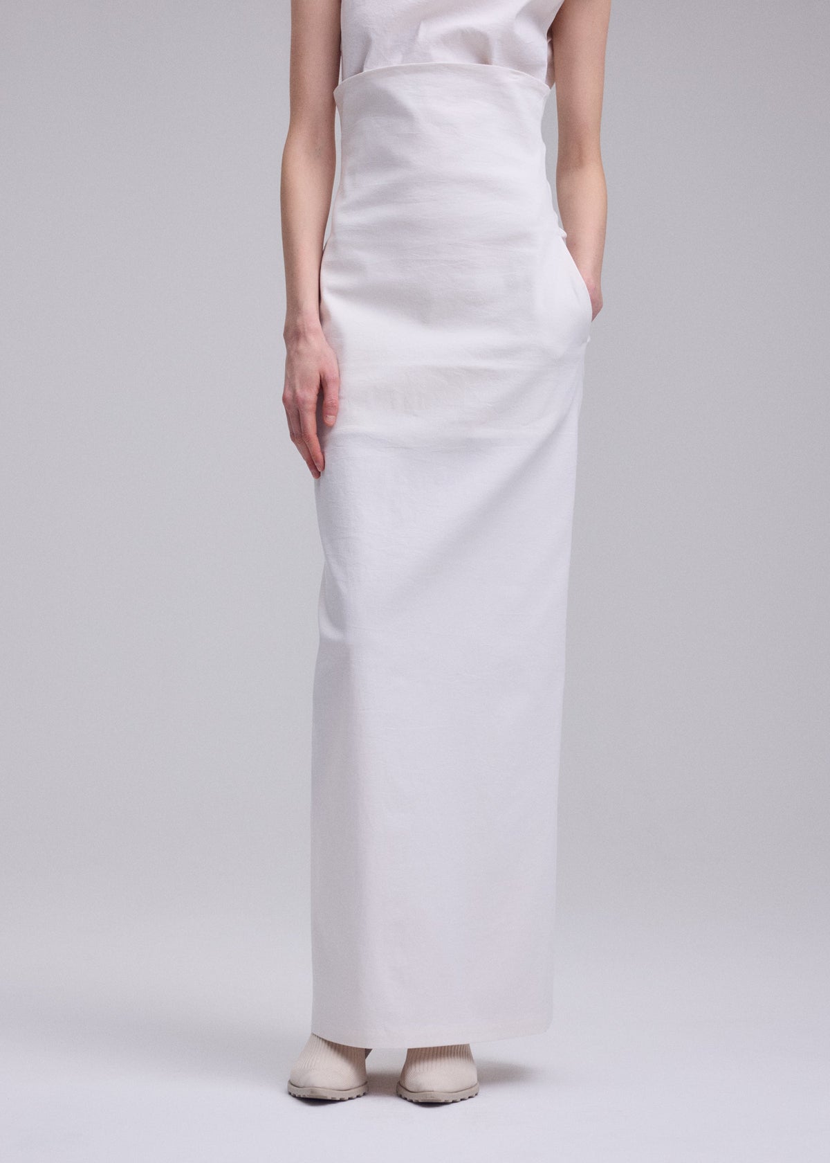 A person wears the ALLOW sleeveless white top and long skirt by ISSEY MIYAKE, standing with hands by their sides against a plain light background. Their face is not visible in the image. view 8 of 9