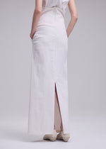 A person stands with hands in pockets, modeling the ALLOW minimalist long white skirt by ISSEY MIYAKE, featuring a back slit and zipper; paired with white shoes, photographed from behind against a plain light background.