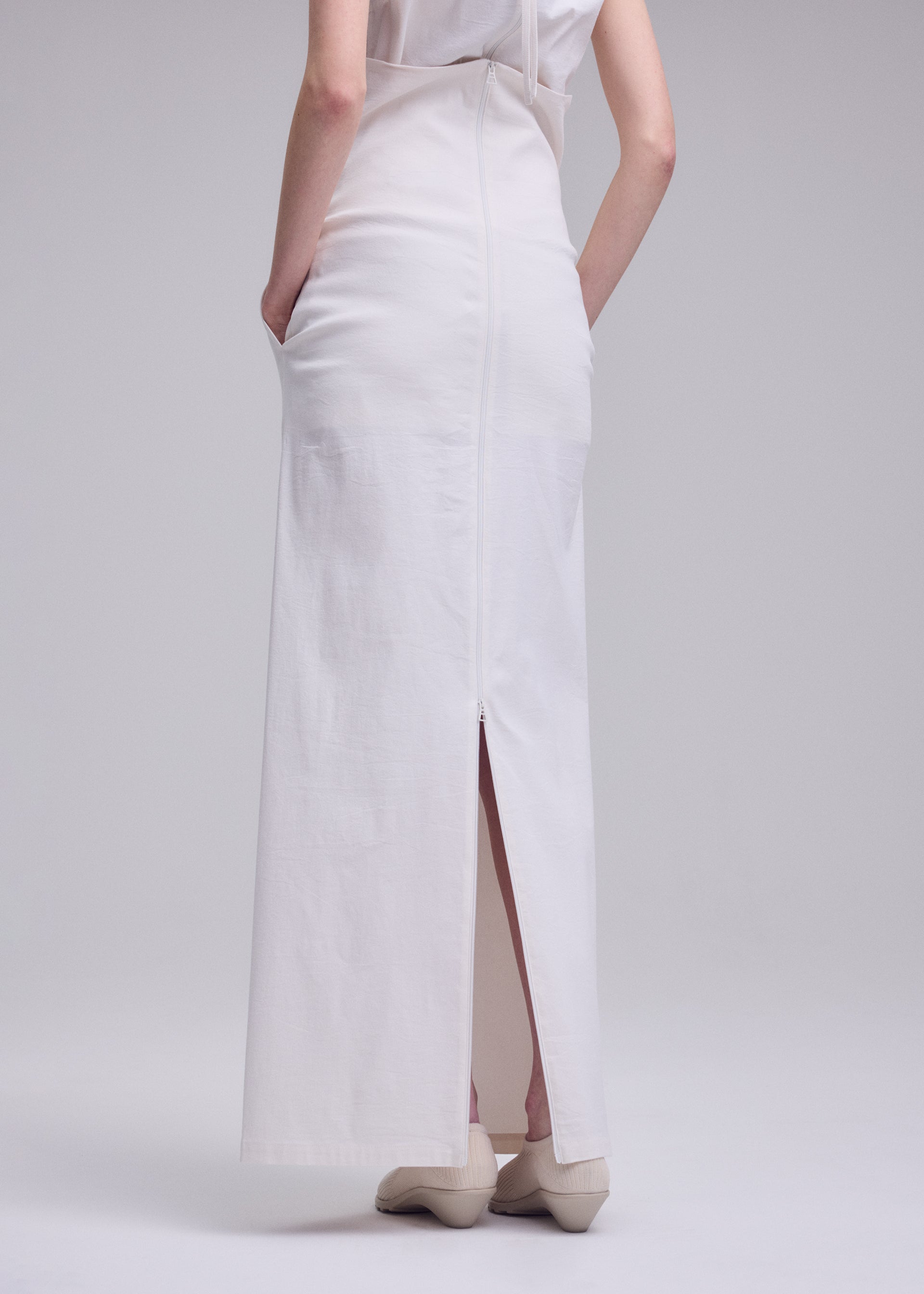 Zoomed view A person stands with hands in pockets, modeling the ALLOW minimalist long white skirt by ISSEY MIYAKE, featuring a back slit and zipper; paired with white shoes, photographed from behind against a plain light background.
