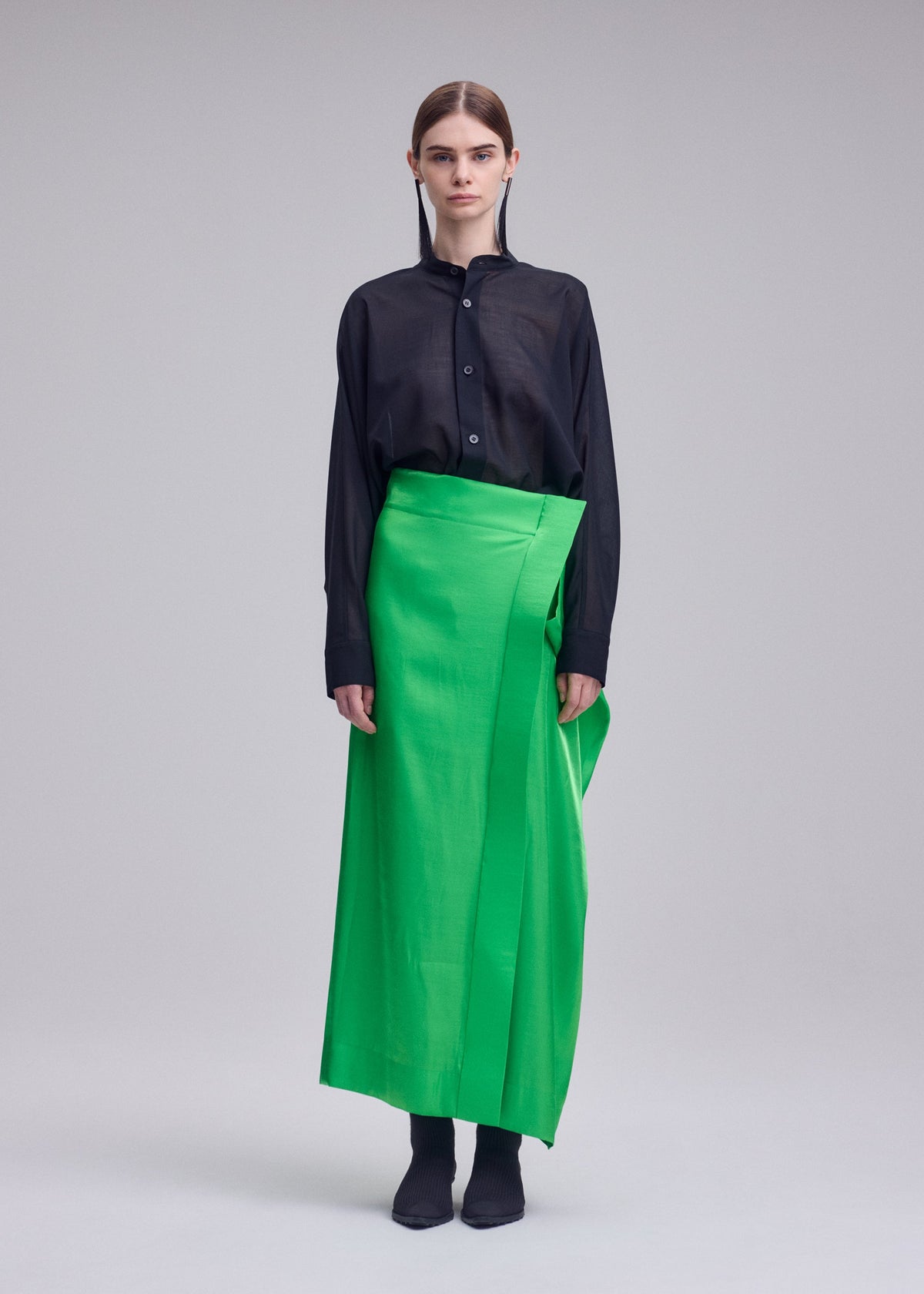 A person stands against a plain background, wearing the ISSEY MIYAKE MERCURY black long-sleeve button-up shirt and a bright green Italian cloth special occasion skirt, paired with black shoes. Their hair is parted in the middle and tucked behind their ears. view 6 of 9