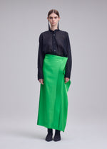 A person stands against a plain background, wearing the ISSEY MIYAKE MERCURY black long-sleeve button-up shirt and a bright green Italian cloth special occasion skirt, paired with black shoes. Their hair is parted in the middle and tucked behind their ears.