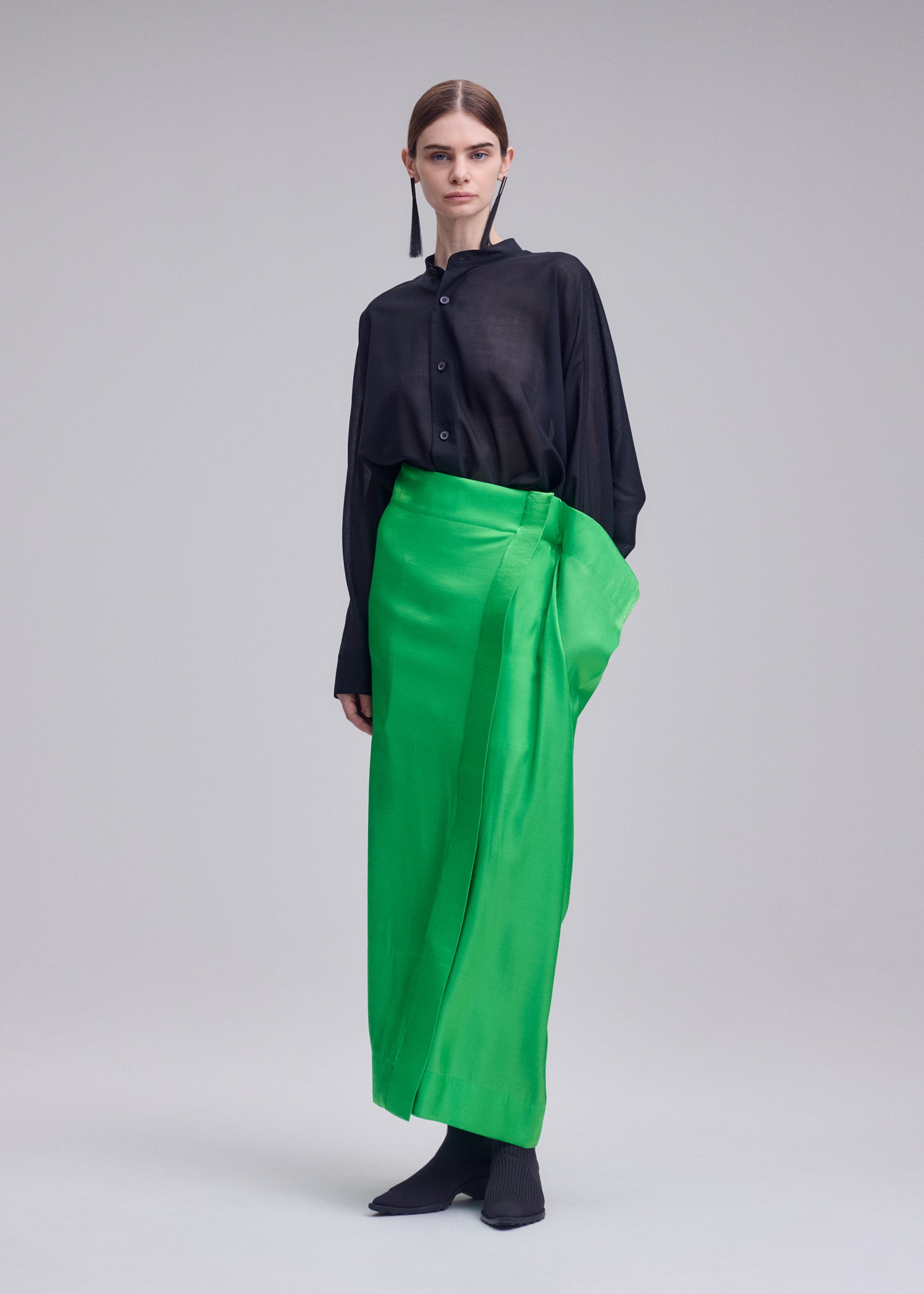 Zoomed view A person wears the ISSEY MIYAKE MERCURY: a loose black long-sleeve blouse and a bright green metallic Italian skirt, paired with black shoes—a standout look ideal for special occasions.