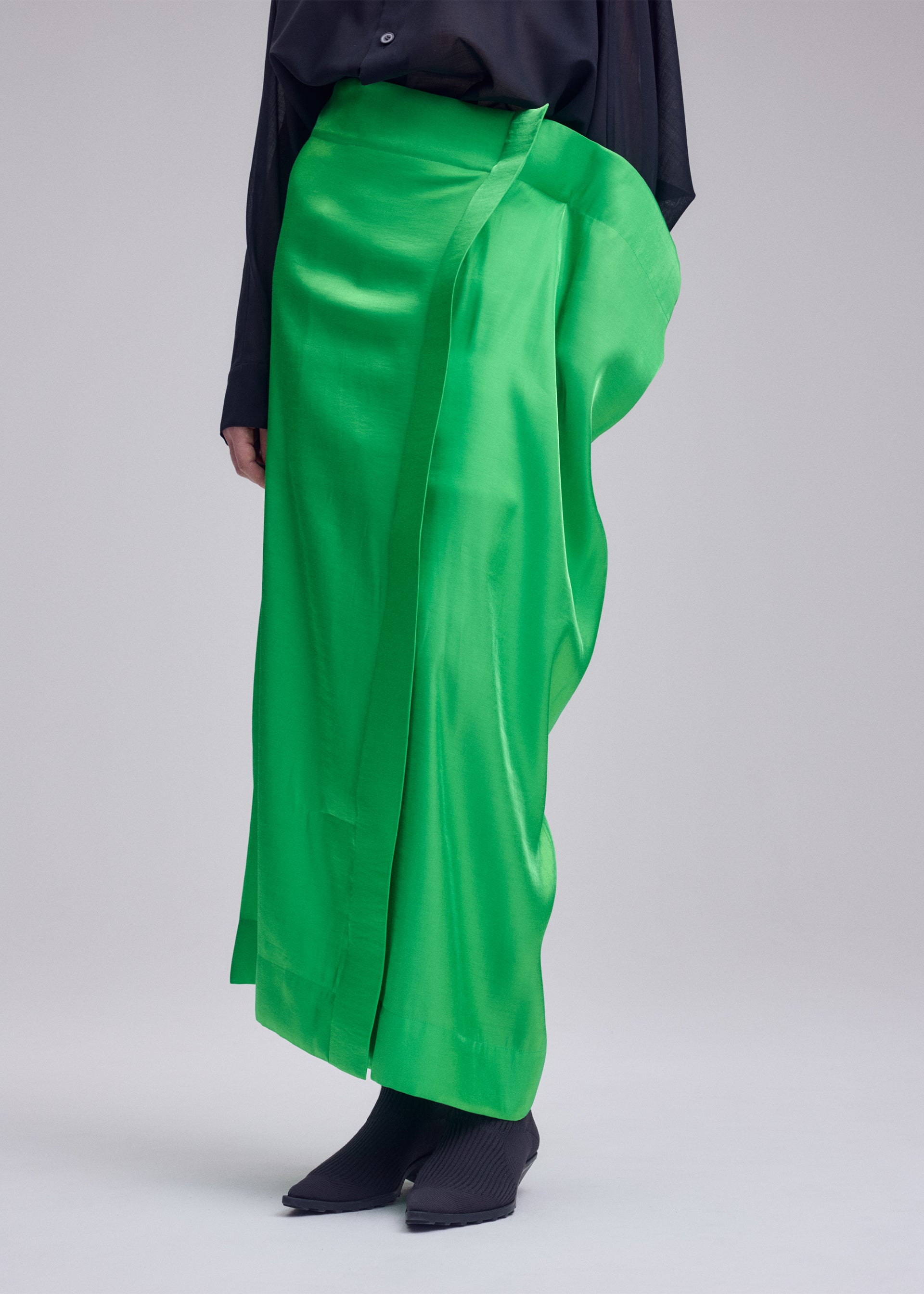 Zoomed view A person wears the shiny, bright green metallic MERCURY skirt by ISSEY MIYAKE, styled with a black long-sleeve top and pointed shoes, standing against a plain light background.