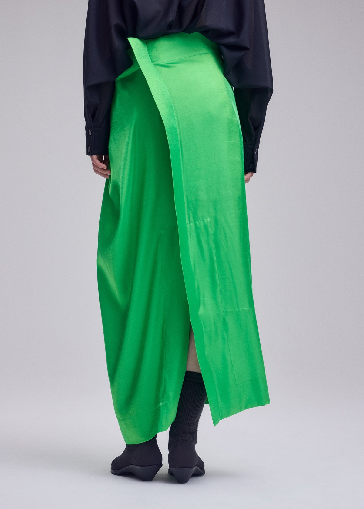 A person stands facing away against a plain background, wearing the ISSEY MIYAKE MERCURY bright green special occasion skirt with a long draped panel, paired with a black long-sleeve top, black socks, and pointed black shoes. view 9 of 9