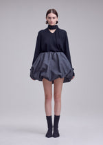 A person stands facing forward, dressed in the ISSEY MIYAKE REFLECTION ensemble—a black long-sleeve top paired with a scarf and a voluminous silver skirt crafted from reflective fabric. They have mid-length hair and wear black socks with black shoes, set against a plain light background.