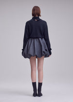 A person stands facing away against a plain light background, wearing ISSEY MIYAKE’s REFLECTION—a dark long-sleeve top paired with a short, puffy reflective silver skirt—and completed with black socks and shoes.