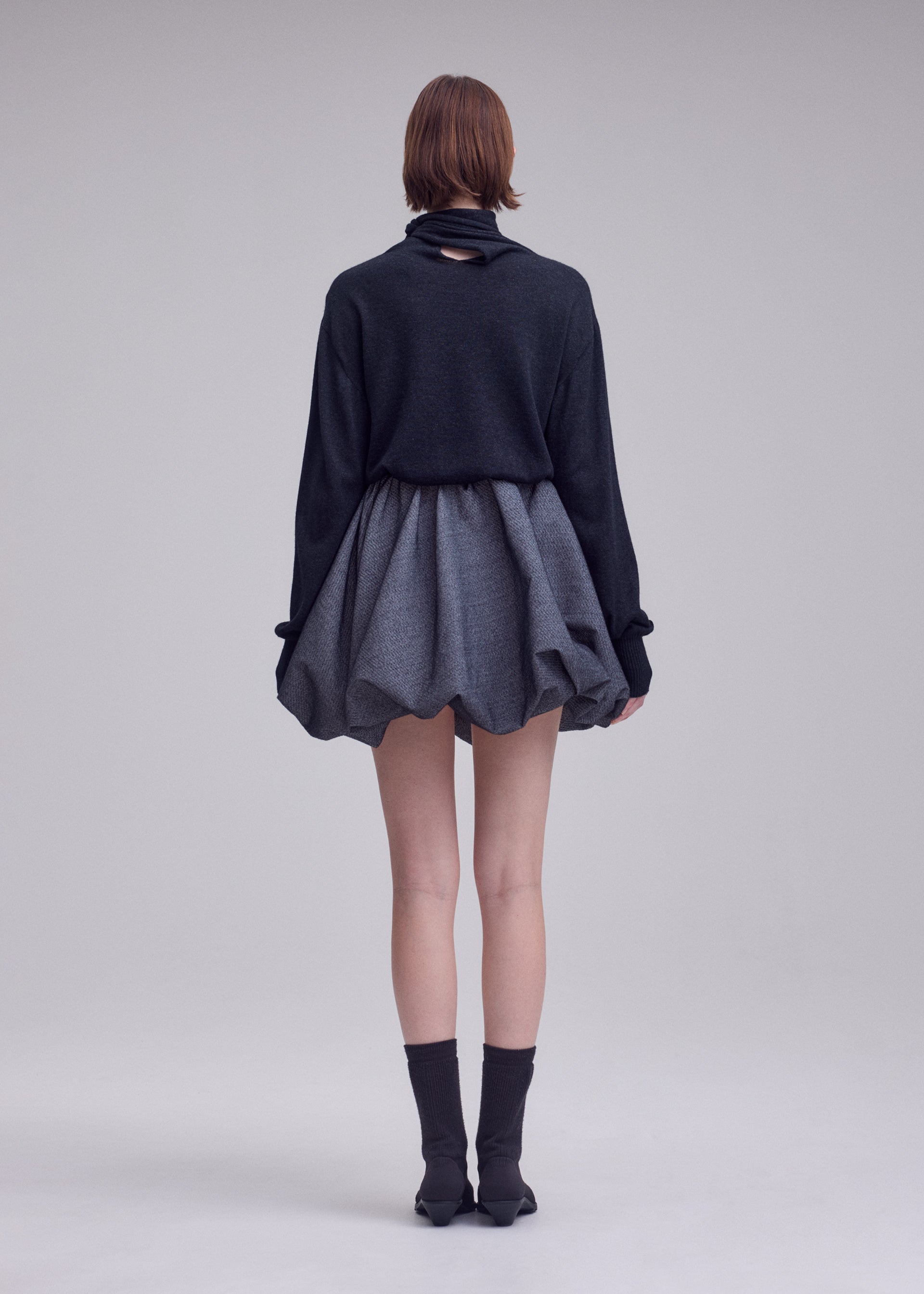 Zoomed view A person stands facing away against a plain light background, wearing ISSEY MIYAKE’s REFLECTION—a dark long-sleeve top paired with a short, puffy reflective silver skirt—and completed with black socks and shoes.
