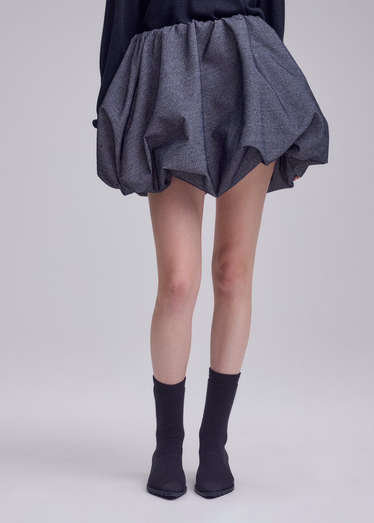 A person stands against a plain light background wearing the ISSEY MIYAKE REFLECTION dark gray reflective skirt, paired with black socks and pointed black shoes. Their upper body is not visible. view 7 of 8