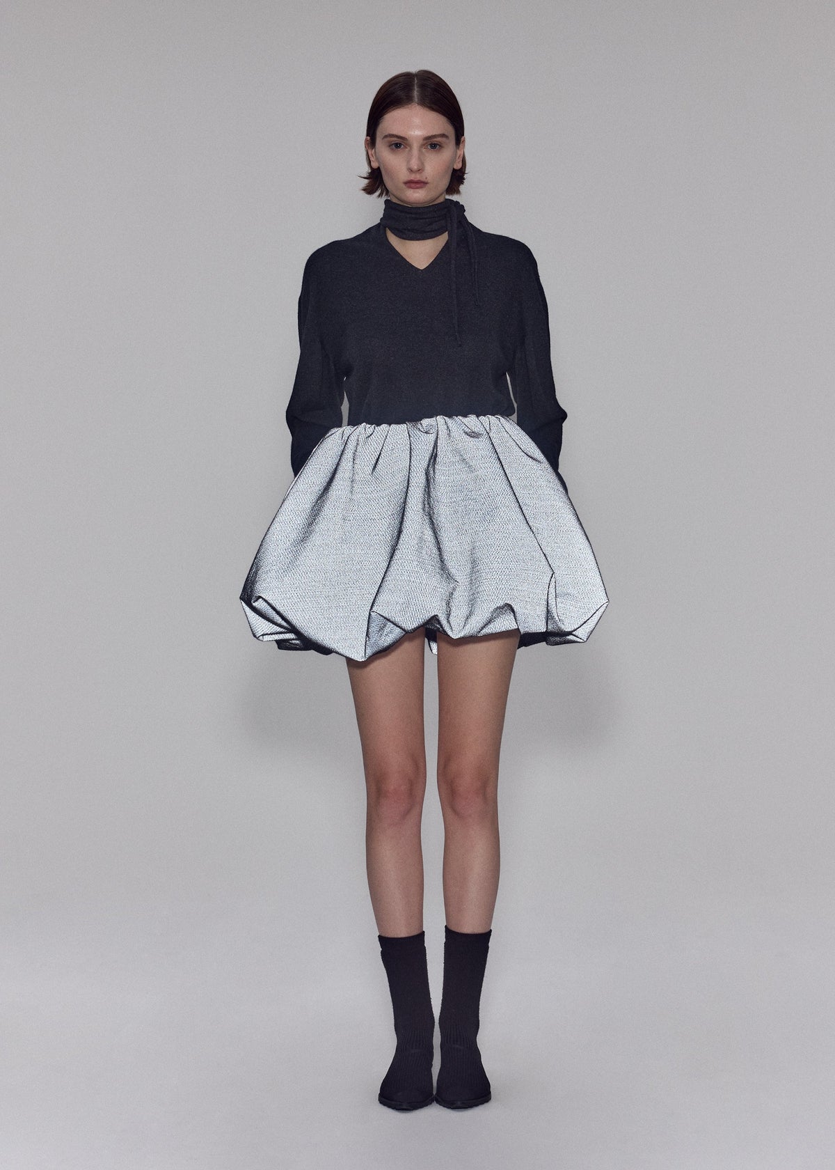A person stands facing forward, wearing the ISSEY MIYAKE REFLECTION dark long-sleeve top with a choker collar and a voluminous silver skirt crafted from reflective fabric. They have short hair, are wearing black socks, and are set against a plain neutral background. view 8 of 8