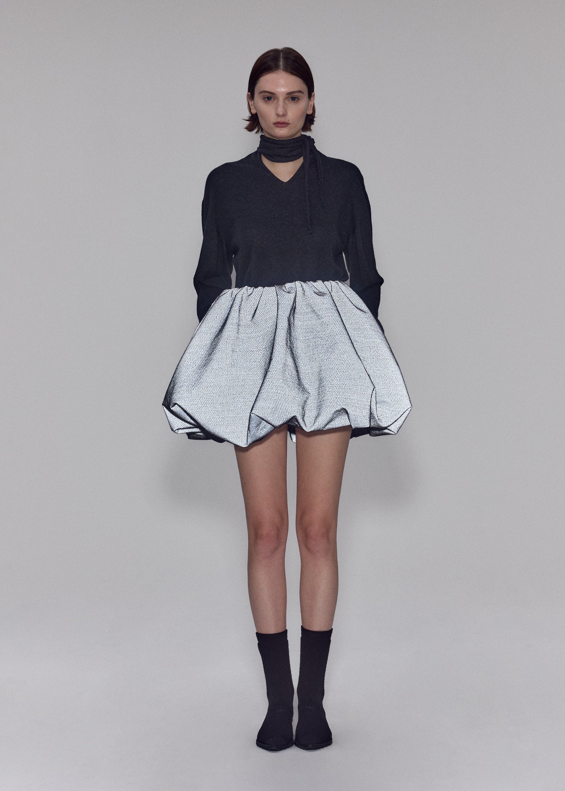 Zoomed view A person stands facing forward, wearing the ISSEY MIYAKE REFLECTION dark long-sleeve top with a choker collar and a voluminous silver skirt crafted from reflective fabric. They have short hair, are wearing black socks, and are set against a plain neutral background.