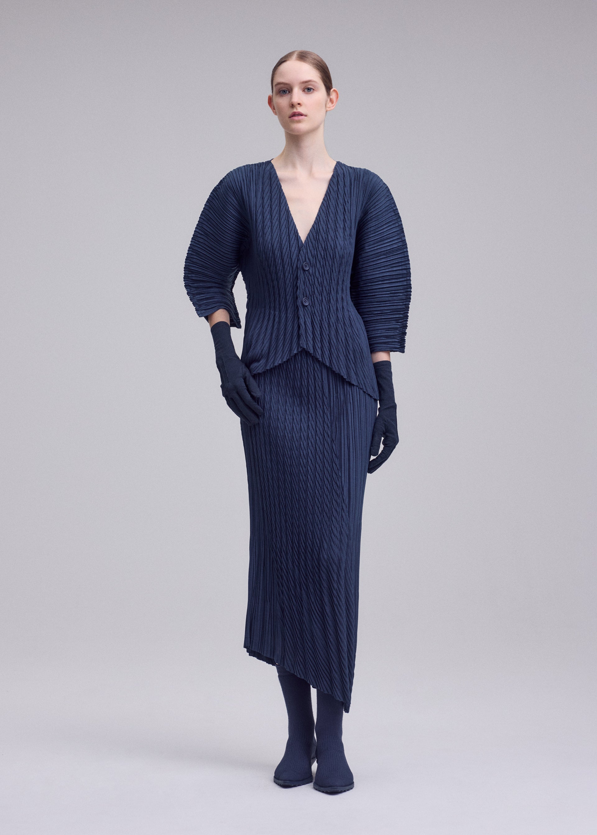Zoomed view A woman poses against a plain backdrop wearing ISSEY MIYAKE’s UNFURLING PLEATS navy skirt and matching three-quarter sleeve top, paired with navy gloves and sock-style shoes, looking directly at the camera with a neutral expression.