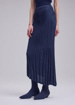 A person wears the UNFURLING PLEATS navy maxi skirt by ISSEY MIYAKE, featuring a slightly sheer finish, paired with matching ribbed socks and pointed-toe shoes against a plain light gray background.
