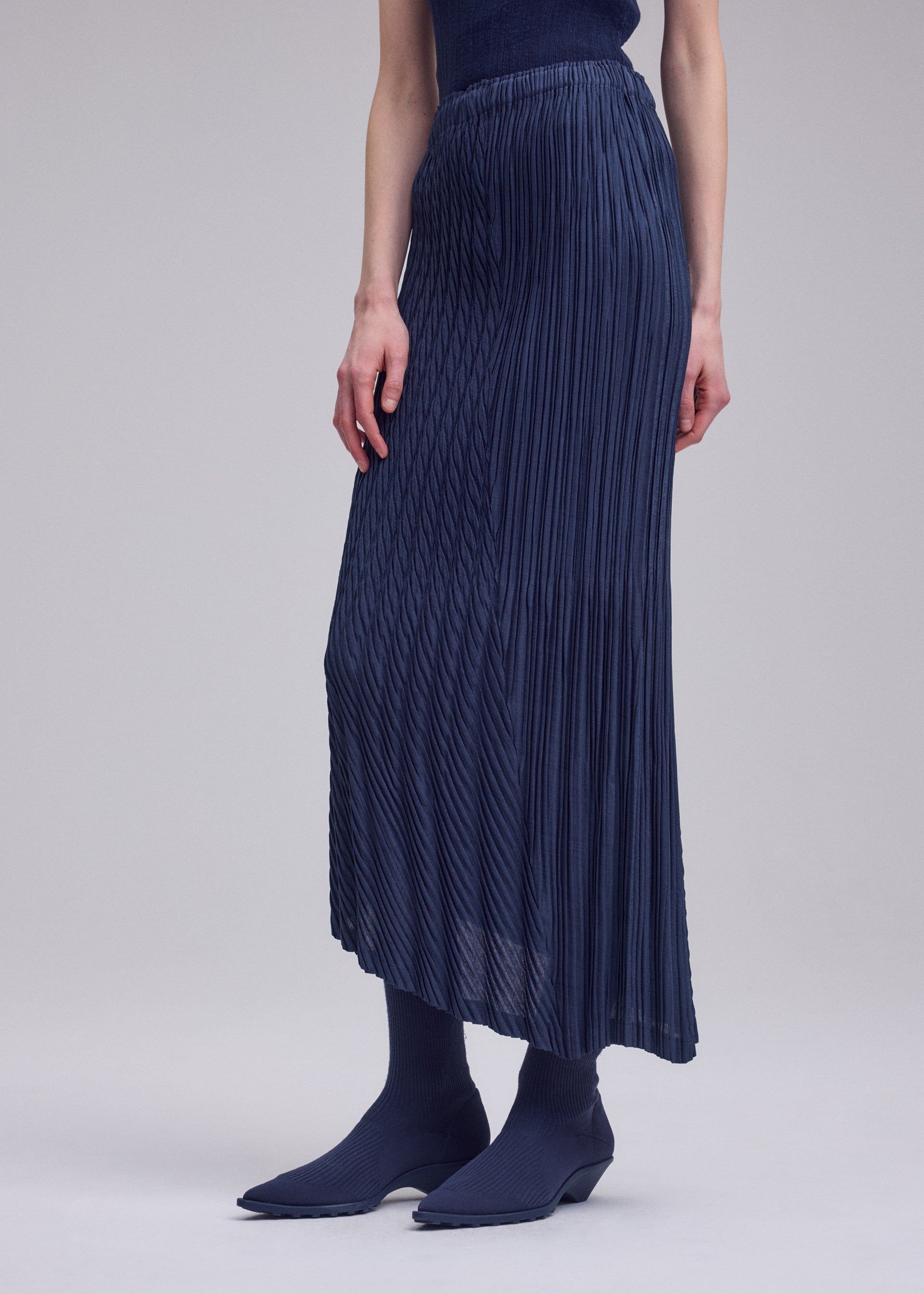 Zoomed view A person wears the UNFURLING PLEATS navy maxi skirt by ISSEY MIYAKE, featuring a slightly sheer finish, paired with matching ribbed socks and pointed-toe shoes against a plain light gray background.
