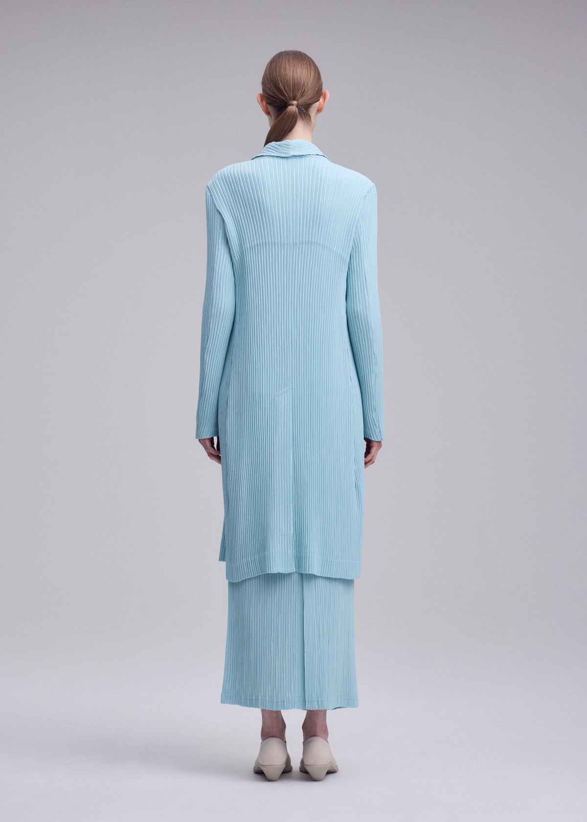 Zoomed view A person with straight, reddish-brown hair wears the ISSEY MIYAKE HATCHING PLEATS long, light blue recycled polyester skirt and matching coat, facing away against a plain gray background with light-colored shoes.