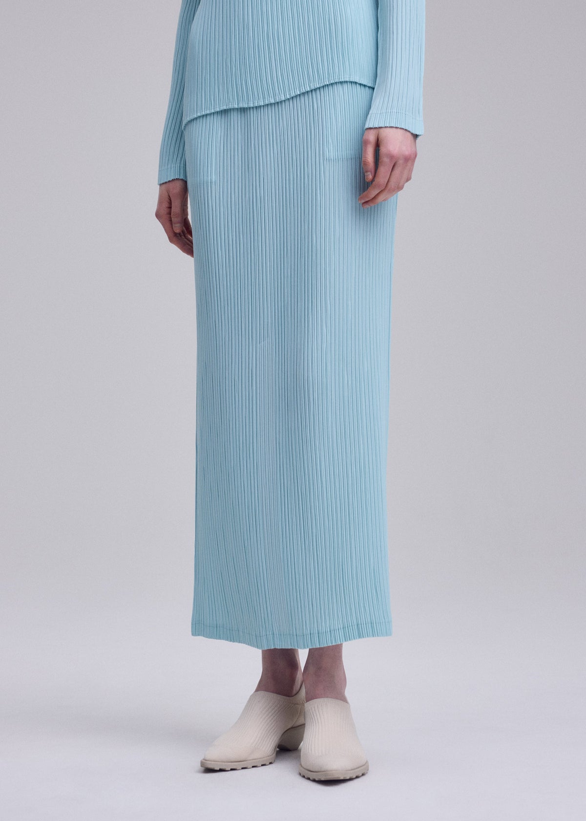 A person wears the ISSEY MIYAKE HATCHING PLEATS light blue, long-sleeve ribbed top with a matching textured pleated skirt and light beige slip-on shoes, standing against a plain background. view 7 of 7
