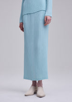 A person wears the ISSEY MIYAKE HATCHING PLEATS light blue, long-sleeve ribbed top with a matching textured pleated skirt and light beige slip-on shoes, standing against a plain background.
