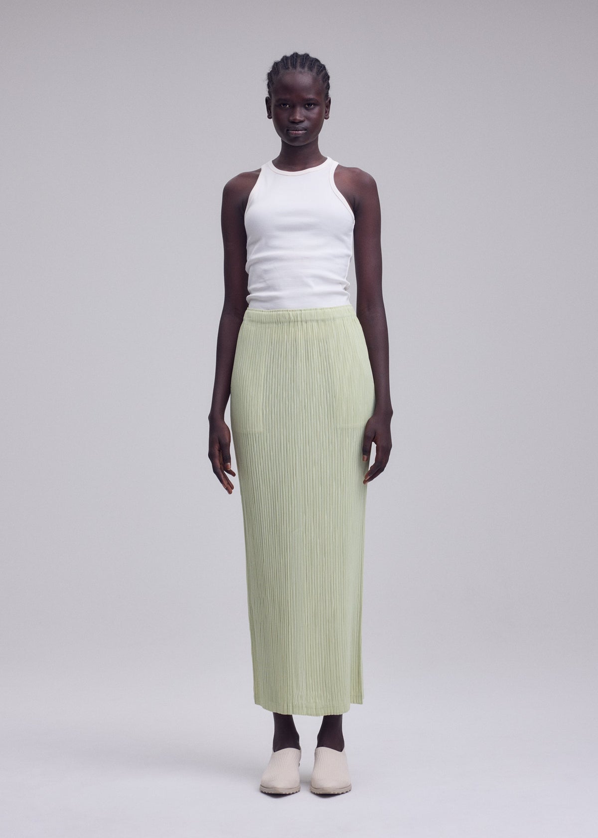 A woman stands against a plain background, wearing a white sleeveless top, beige shoes, and the ISSEY MIYAKE HATCHING BOTTOMS—a light green long pleated skirt with a subtle textured finish. view 5 of 8
