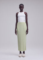 A woman stands against a plain background, wearing a white sleeveless top, beige shoes, and the ISSEY MIYAKE HATCHING BOTTOMS—a light green long pleated skirt with a subtle textured finish.