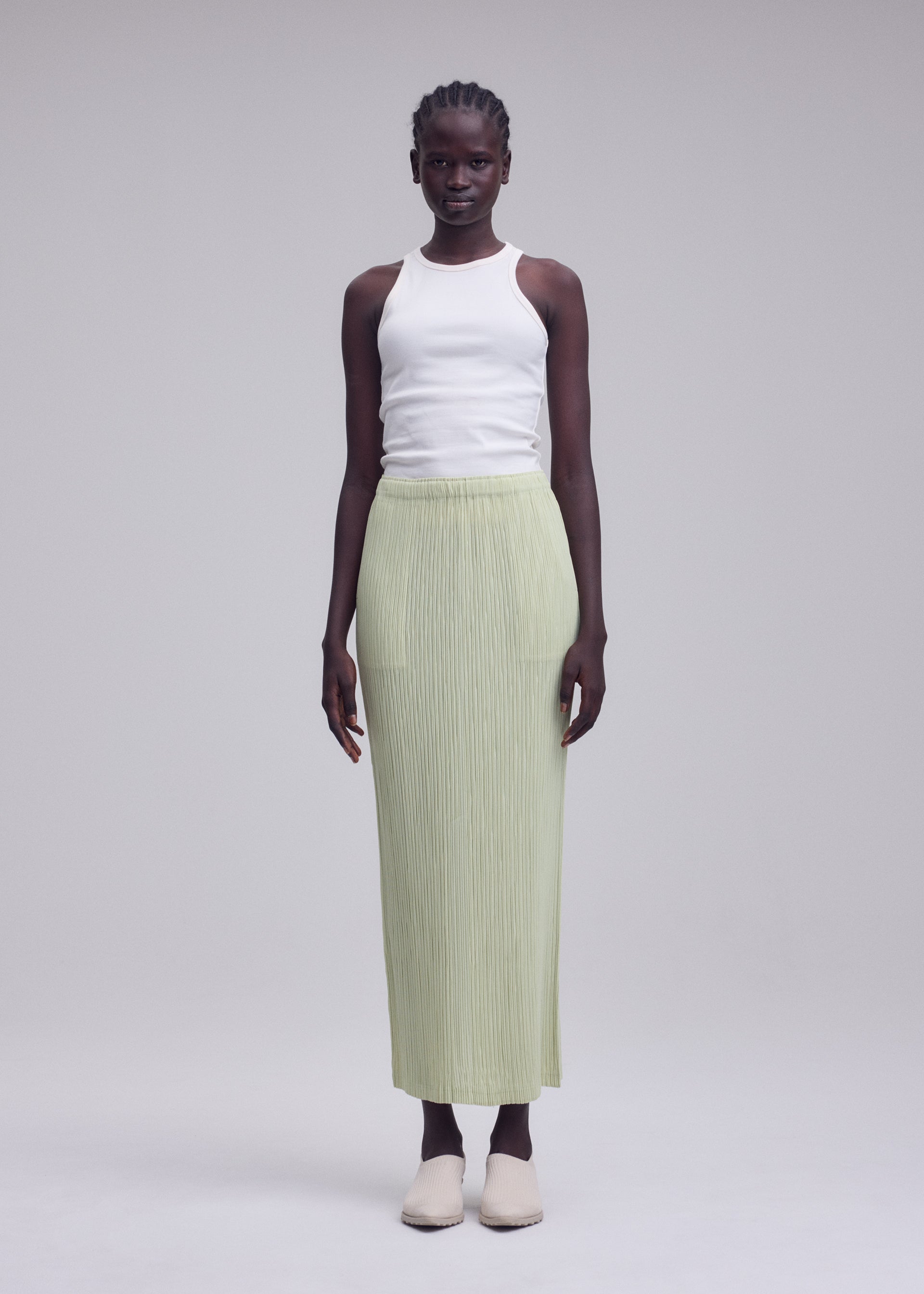 Zoomed view A woman stands against a plain background, wearing a white sleeveless top, beige shoes, and the ISSEY MIYAKE HATCHING BOTTOMS—a light green long pleated skirt with a subtle textured finish.