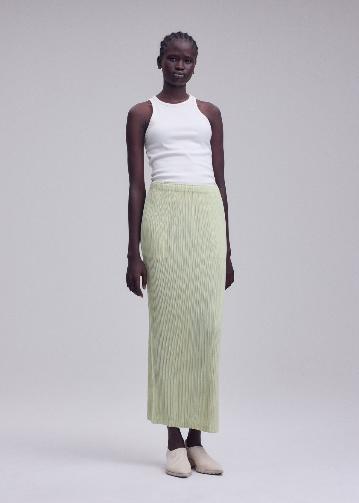 A woman stands against a plain background wearing a white sleeveless top, beige shoes, and the ISSEY MIYAKE HATCHING BOTTOMS—a light green, long pleated polyester skirt with a textured finish. view 6 of 8