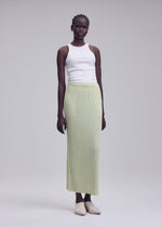 A woman stands against a plain background wearing a white sleeveless top, beige shoes, and the ISSEY MIYAKE HATCHING BOTTOMS—a light green, long pleated polyester skirt with a textured finish.