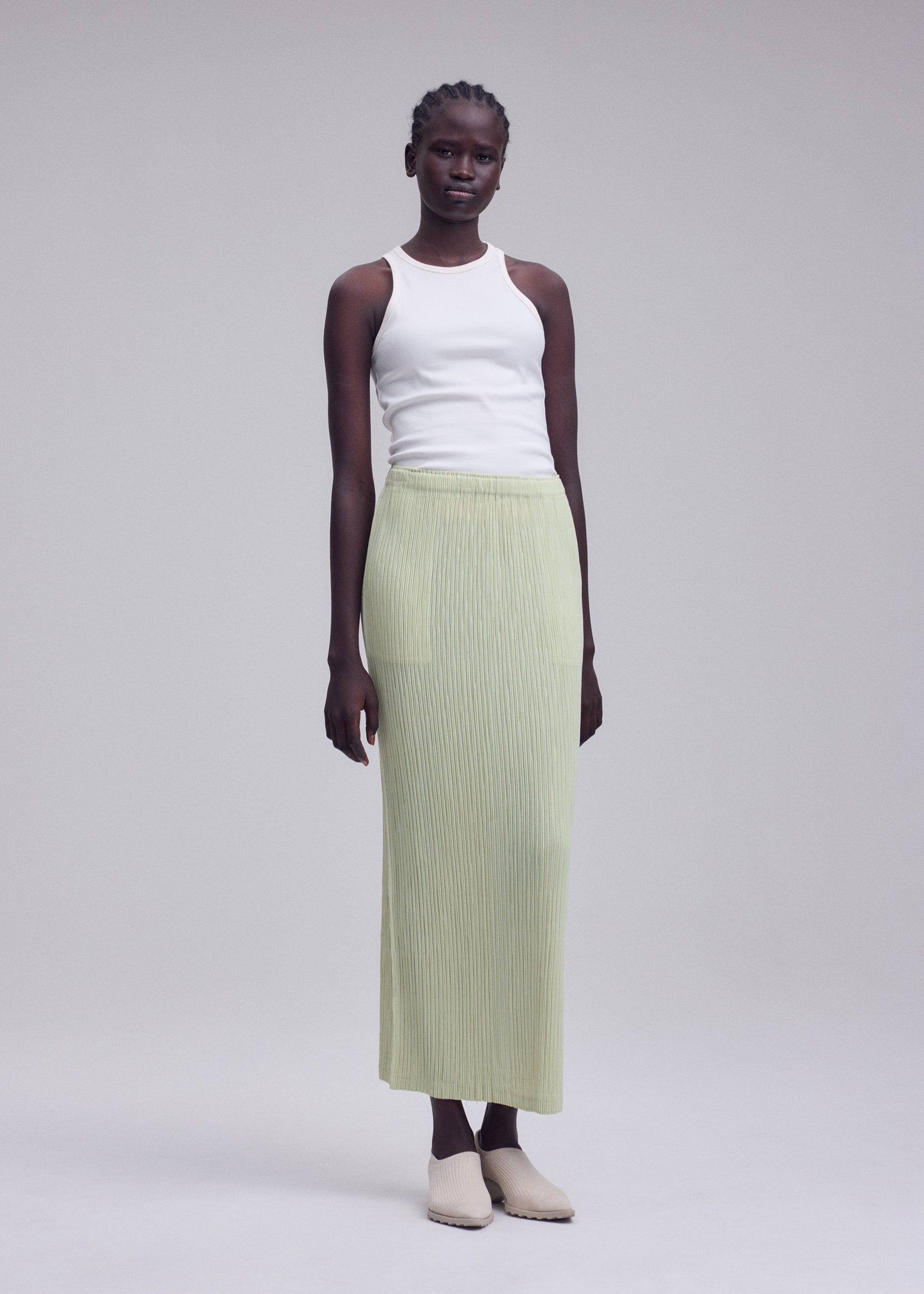 Zoomed view A woman stands against a plain background wearing a white sleeveless top, beige shoes, and the ISSEY MIYAKE HATCHING BOTTOMS—a light green, long pleated polyester skirt with a textured finish.