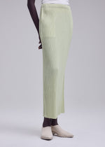 Dressed in ISSEY MIYAKE’s HATCHING BOTTOMS, a person stands against a plain light background, paired with a white top and beige slip-on shoes. The photo is cropped from shoulder to feet.