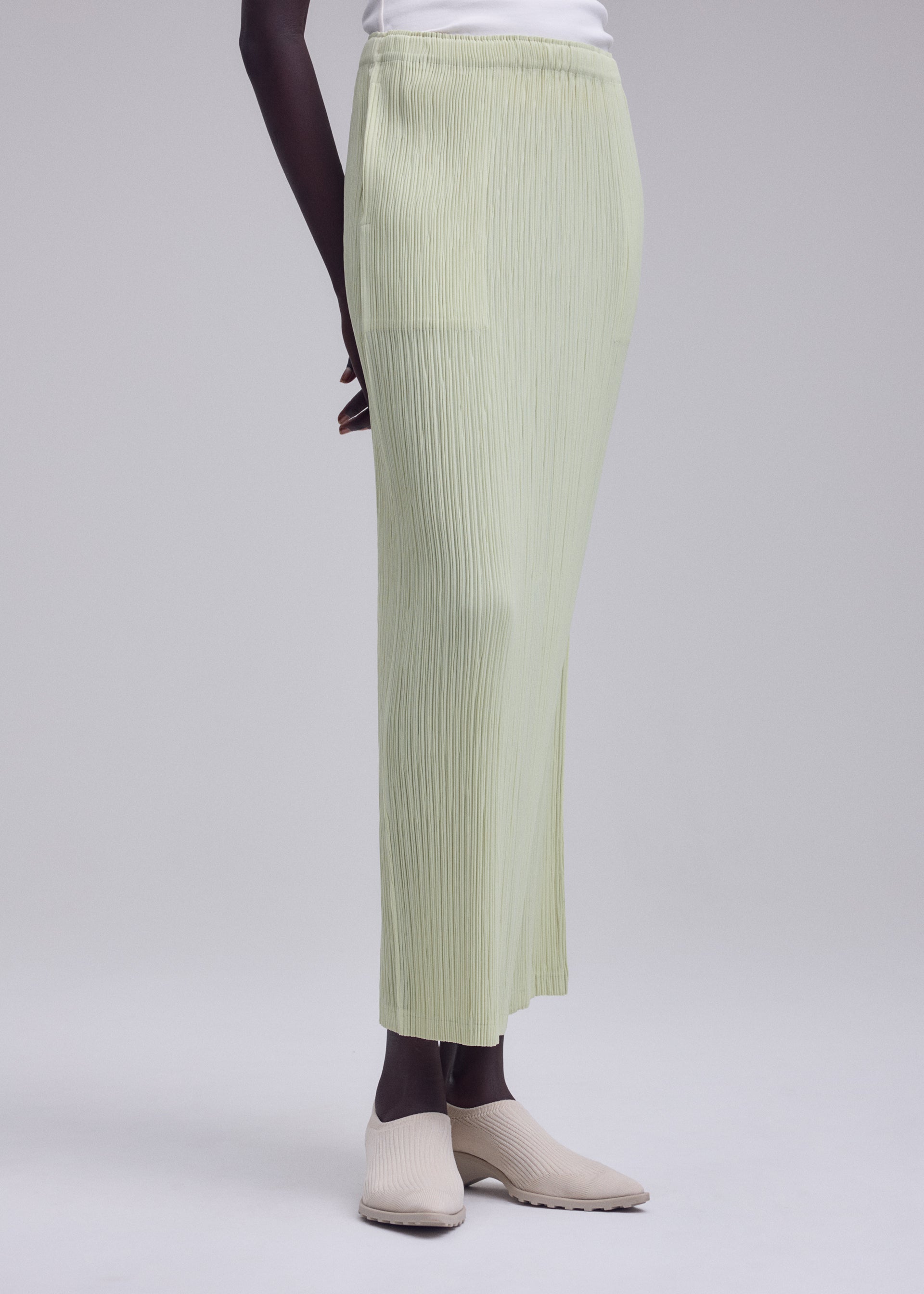 Zoomed view Dressed in ISSEY MIYAKE’s HATCHING BOTTOMS, a person stands against a plain light background, paired with a white top and beige slip-on shoes. The photo is cropped from shoulder to feet.