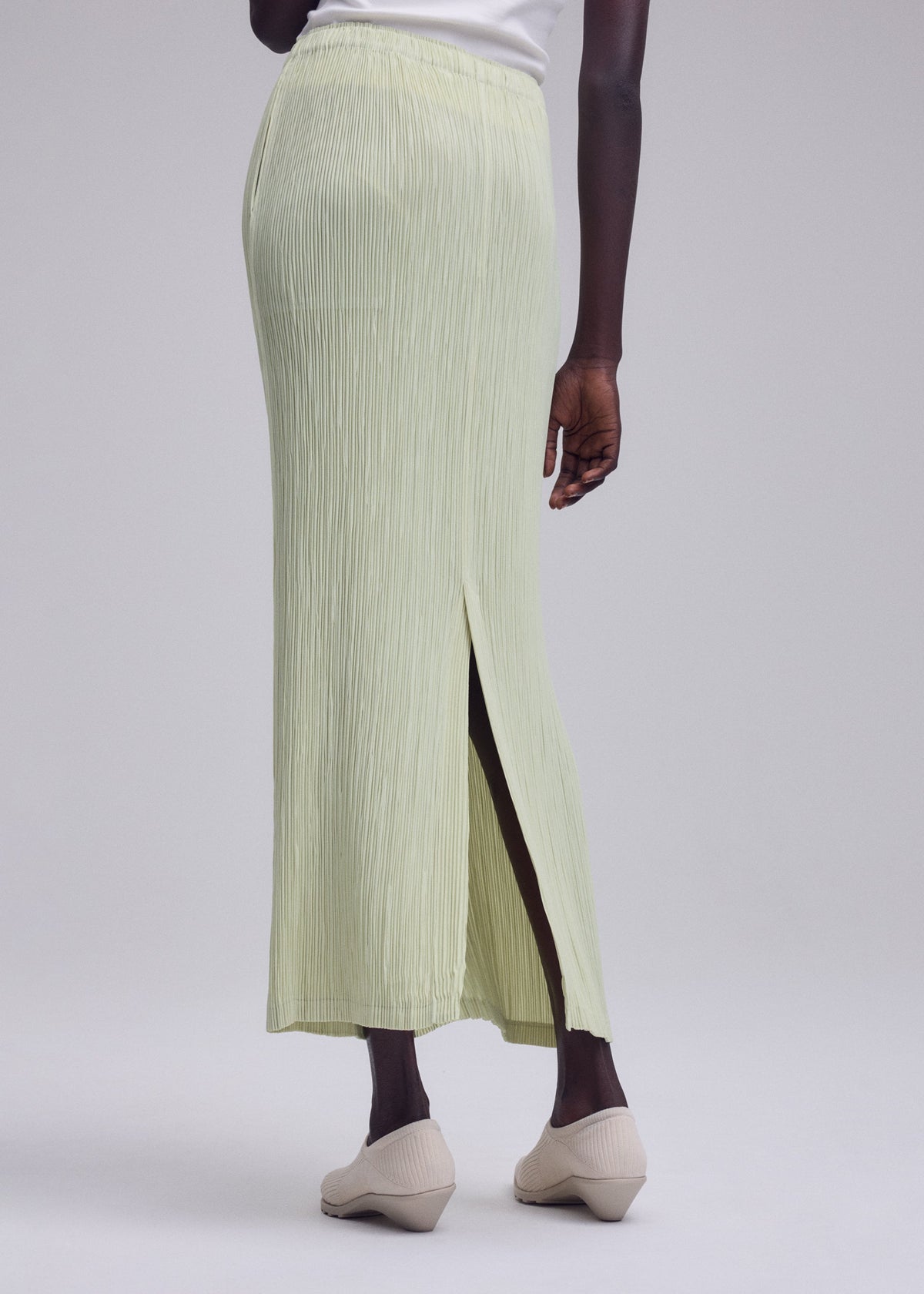 A person is standing against a plain light gray background, dressed in the ISSEY MIYAKE HATCHING BOTTOMS—a light green, long pleated skirt with a textured finish and back slit—paired with beige shoes and a white top. view 8 of 8