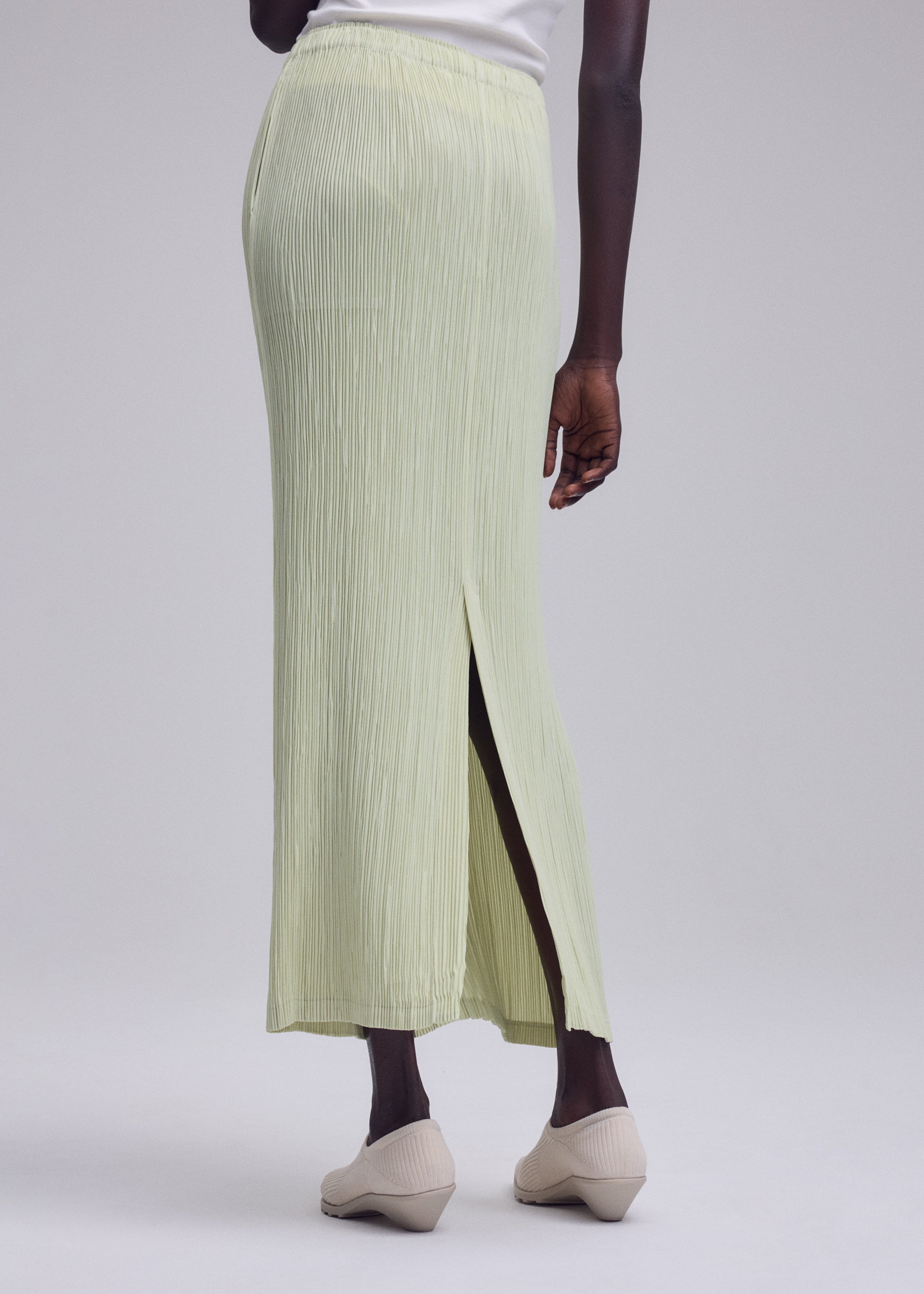 Zoomed view A person is standing against a plain light gray background, dressed in the ISSEY MIYAKE HATCHING BOTTOMS—a light green, long pleated skirt with a textured finish and back slit—paired with beige shoes and a white top.
