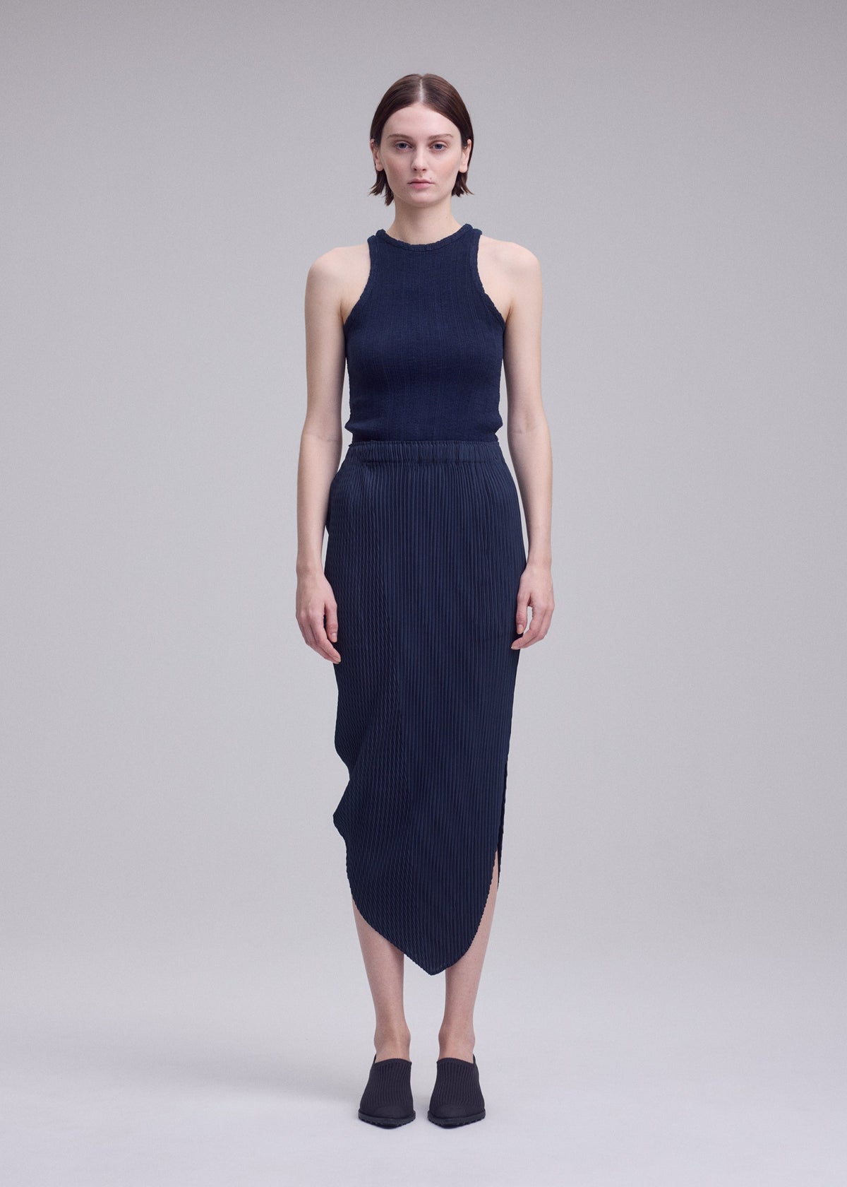 A woman with short hair and a neutral expression stands against a plain background, wearing the ISSEY MIYAKE LEAF PLEATS set—a sleeveless, dark ribbed top paired with a matching leaf silhouette skirt—and black slip-on shoes. view 6 of 8