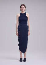 A woman with short hair and a neutral expression stands against a plain background, wearing the ISSEY MIYAKE LEAF PLEATS set—a sleeveless, dark ribbed top paired with a matching leaf silhouette skirt—and black slip-on shoes.